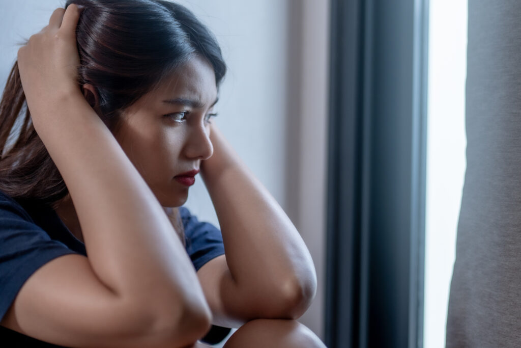 Una mujer sentada junto a una ventana, de aspecto pensativo y angustiado, con las manos en la cabeza. la luz suave crea una atmósfera contemplativa