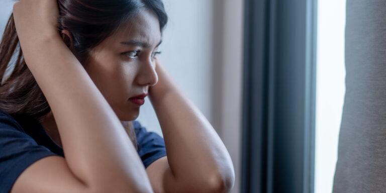 A woman sitting by window, looking pensive and distressed, with her hands on her head. soft light creates contemplative atmosphere